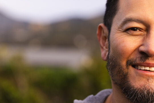 Asian man smiling while standing by lake with blurred shrubs and hills in hoodie, copy space