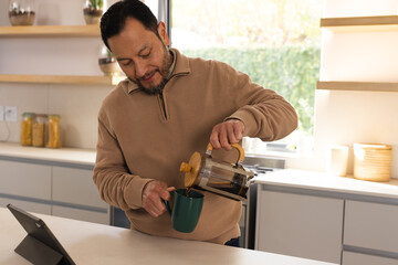 Asian man wearing pullover at kitchen island pouring coffee from French press into mug with tablet