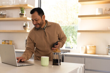 Mature Asian man making coffee with French press on kitchen island typing on laptop with smartphone