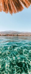 Crystal-clear turquoise sea with visible underwater rocks, distant coastline, and a straw beach umbrella overhead against a clear blue sky.