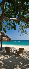 Two wooden chairs and a table sit under a tree on a sandy beach, overlooking turquoise water with boats in the distance under a clear blue sky.