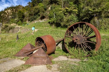 A girl hikes past historic gold mining equipment in Karangahake Gorge, New Zealand. The rusty...