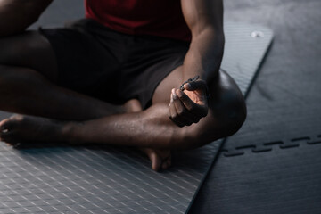Mature African American man in sportswear sitting on mat over foam tiles at gym holding mudra