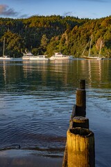 Boats are moored in a calm bay in New Zealand. The wooden posts of a pier are in the foreground,...