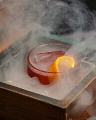 A smoky, dark red alcoholic cocktail with ice and an orange peel garnish is presented in a rocks...