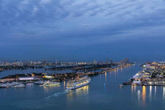 Aerial view of a cruise ship glides through the shimmering waterway, surrounded by the city lights and the vastness of the ocean, Miami, Florida, United States.