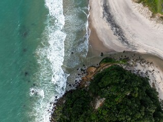Aerial view of waves crashing on a sandy beach and rocky coastline covered in lush green trees. The...