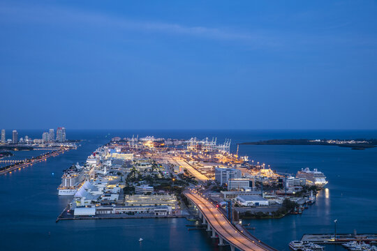 Aerial view of Dodge Island glows with warm lights against the deep blue of the surrounding water and sky, connected by a bridge bustling with activity, Miami, Florida, United States.