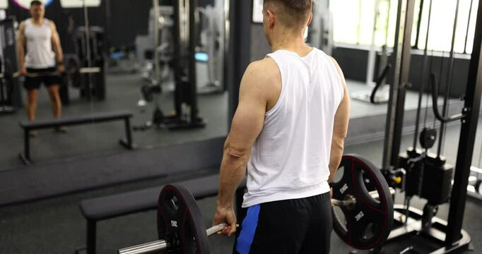 Strong man in activewear lifts barbell with shoulders in gym backside view. Skilled bodybuilder trains muscles working with large weight near mirror