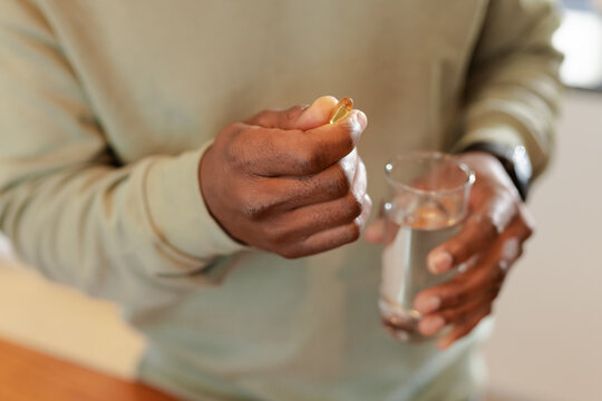 African American man holding glass of water and pinching softgel capsule at home kitchen counter