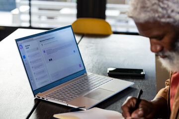 Senior African American man jotting notes at office desk beside laptop, phone by window, copy space