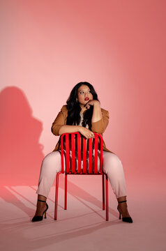 Female model posing astride red metal chair in photography studio with pink gradient backdrop
