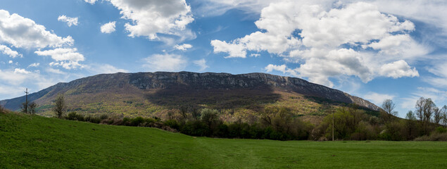 Amazing panorama of a green meadow in the mountains. The meadow is covered with a blue sky filled with white fluffy clouds. A huge mountain is visible in the background. Spring or summer nature photo.