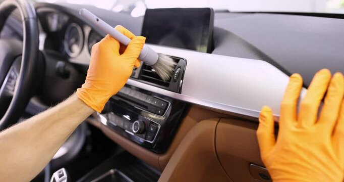 Person in orange gloves meticulously cleans interior of car using brush to dust air vents on dashboard. Process of car detailing and maintenance