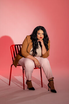 Woman sitting on red metal chair in studio with pink gradient backdrop wearing brown blazer
