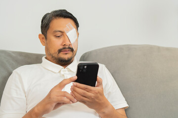 A bearded man with white medicine plaster patch on the eye, sits on a cloth sofa