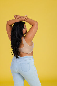 Woman raising arms above head and turning head against yellow backdrop wearing sports bra and jeans