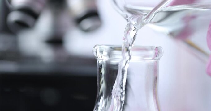 Chemist pours transparent liquid from jar into tall glass container. Light passes through water creating sparkling highlights on surface