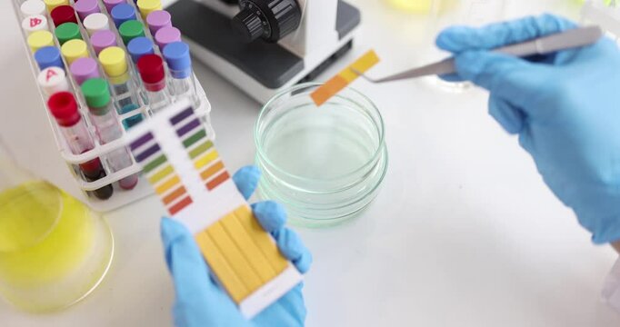 Technician in blue gloves dips pH strip into liquid testing with color chart. Bright tubes and Petri dish reflect organized scientific lab setting