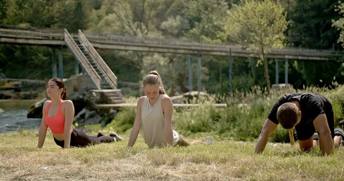 Three friends practice yoga poses in nature by the river. They perform a cobra pose and transition to a cat pose.