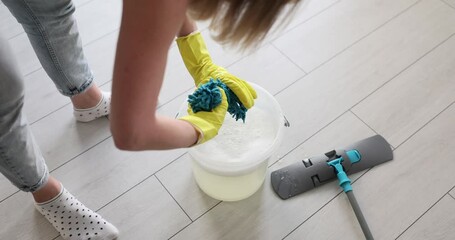 Woman in yellow gloves wrings out blue mop head over clear bucket filled with water. Grey mop on tiled floor near bucket indicates cleaning task