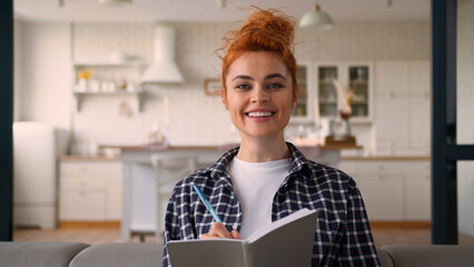 Smiling happy Caucasian woman girl lady designer homeowner owner own apartment writing ideas in notebook noting write information schedule checklist manage budget smile at camera female at home couch