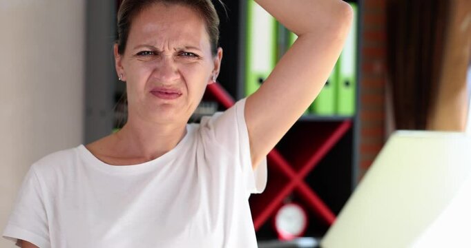 Adult woman sniffs underarm grimacing in disgust from strong odor. Facial reaction reveals sudden realization of bad smell after work day in office