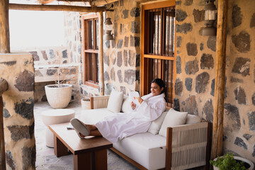 A woman in a robe relaxes on a patio sofa reading a book in a heritage resort The stone walls and...