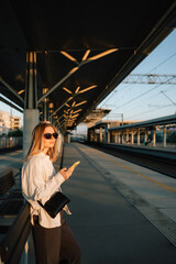 Woman waiting for train at railway station during sunset, using her smartphone to check schedules...