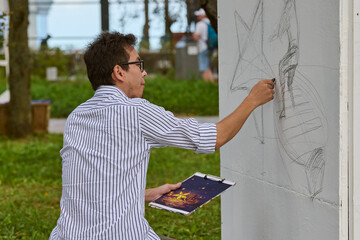 Young asian male artist sketching with pencil on outdoor wall. Focused expression, striped shirt,...