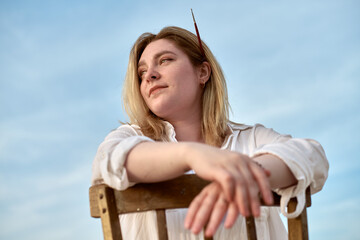 Young woman gazes thoughtfully under clear sky, leaning on wooden chair. With paintbrush in her...