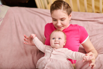 Young female with light brown hair and baby girl in bright room. Mother in pink top, daughter in...