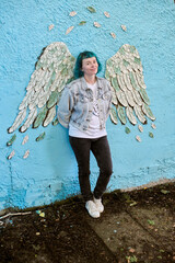 Young woman with blue hair stands against turquoise wall with painted angel wings. Denim jacket and...