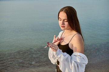 Young adult female painter stands by calm lake, holding paintbrush. Her expression reflects...