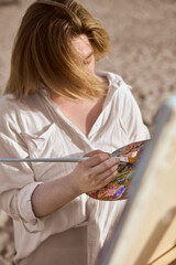 Young female artist paints with vibrant colors on palette in natural light. She stands outdoors,...