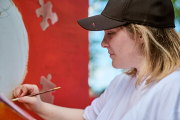 Young caucasian female painting vibrant mural with brush, daylight illuminating artwork. Red,...