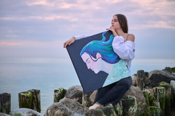 Young female painter sits on rocky shores at sunset, holding vibrant portrait. Serene coastal...