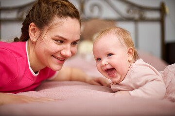 Young woman with brown hair and smiling baby girl on pink bed. Light-hearted interaction, creating joyful atmosphere. Warm lighting enhances bond