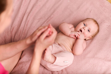 Young mother with light hair assists infant girl with passive exercises. Comfortable indoor setting...