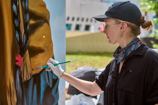 Young adult caucasian female artist in black shirt and cap painting mural outdoors. Woman focused, wearing sunglasses, surrounded by greenery, vibrant colors, natural light - Powered by Adobe