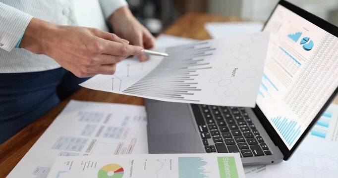 Person extends document sheet toward laptop covered with charts and stats. Hand reaches over table creating scene of data-driven presentation