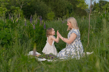 A happy mom and little daughter are sitting on a blanket in the park, clapping their hands. A happy...
