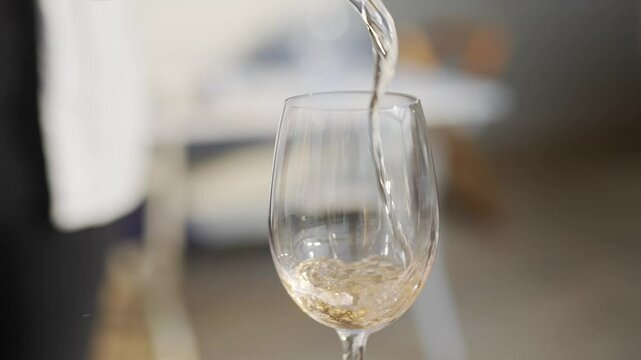 A person is pouring wine into a wine glass. The wine glass is half full and the wine is being poured into the glass