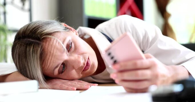 Exhausted woman in lab coat puts head on table falling asleep while holding pink smartphone. Medical professional experiences burnout in clinic