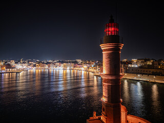 Aerial view of the illuminated Lighthouse of Chania standing tall against the dark sky, with city lights reflecting in the calm waters, Chania, Chania, Greece.