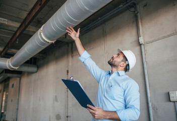 Confident construction engineer in hard hat inspecting industrial pipes indoors, holding a clipboard, demonstrating professional assessment, project management, and safety protocols in a building stru