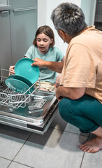 Child helps grandmother to load dishwasher. Family everyday life concept.