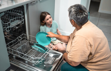 Child helps grandmother to load dishwasher. Family everyday life concept.