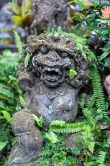 Stone statue of a Balinese demon, a protective figure, covered in moss and ferns, in a garden in Ubud, Bali, Indonesia. The statue is weathered and old.