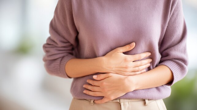A woman holding her stomach in pain from discomfort. A person in a pale purple sweater and beige pants experiencing bloating. Hands on a belly due to digestive disorder